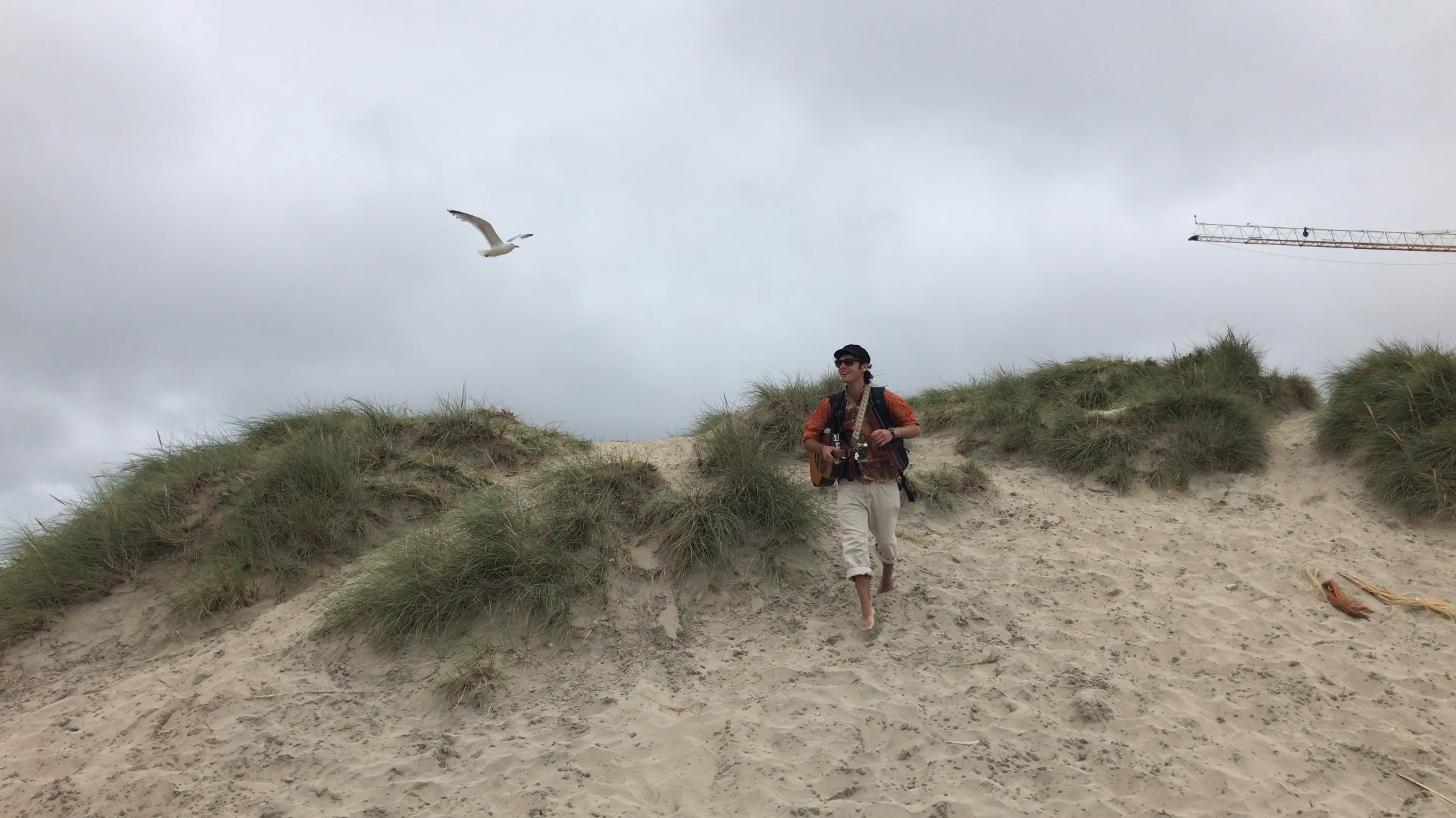 Albert Romiel walking down the sand dunes at Perranporth, Cornwall, guitar on his back, seagull overhead